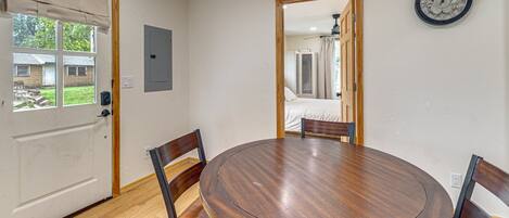 Un comedor con una mesa redonda de madera y cuatro sillas, un reloj en la pared y una puerta que da a otra habitación.