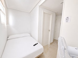 Una habitación pequeña con cama individual, ropa de cama blanca, un mando a distancia sobre la cama, una puerta a la derecha y un cartel de Wi-Fi en la pared.