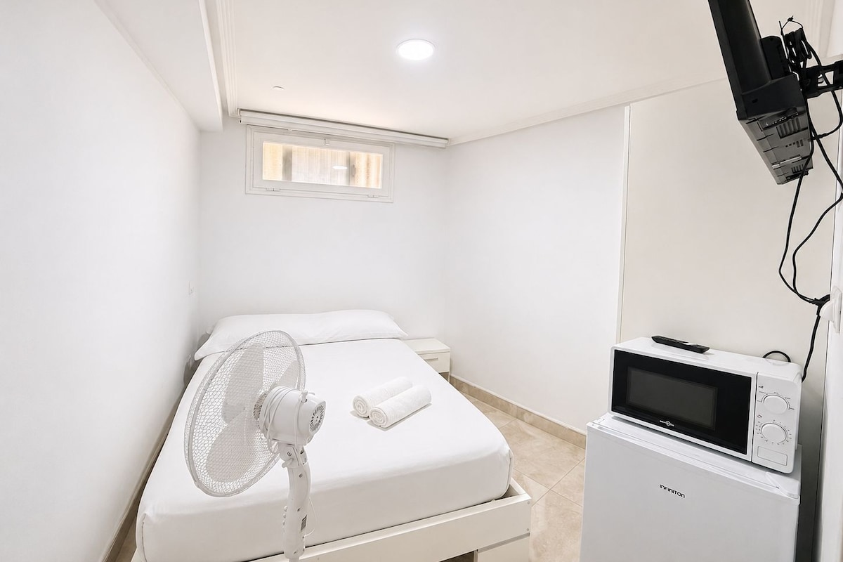 Un dormitorio con cama, ventilador, microondas y un calefactor pequeño.