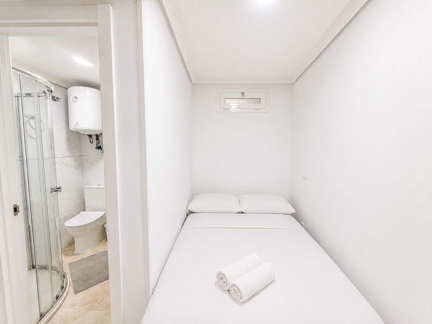 A small, white, single-room hotel bathroom with a shower, toilet, and a bed with rolled towels.