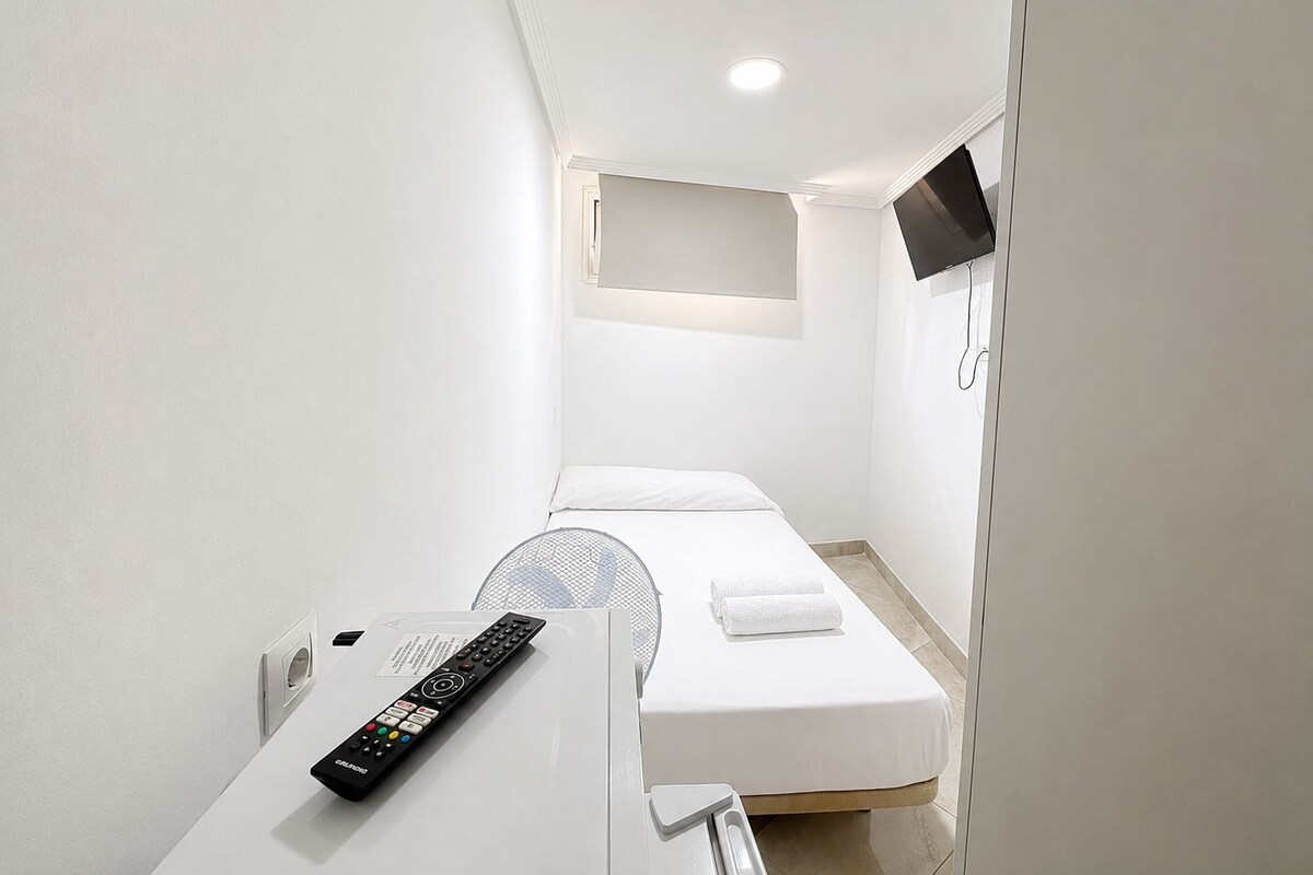 Habitación de hotel compacta con una cama individual, una mesita de noche con un control remoto, un televisor montado en la pared y una ventana pequeña.