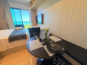 Habitació d'hotel amb un llit, una taula de dinar preparada amb coberts, un vas amb flors i un televisor penjat a la paret.