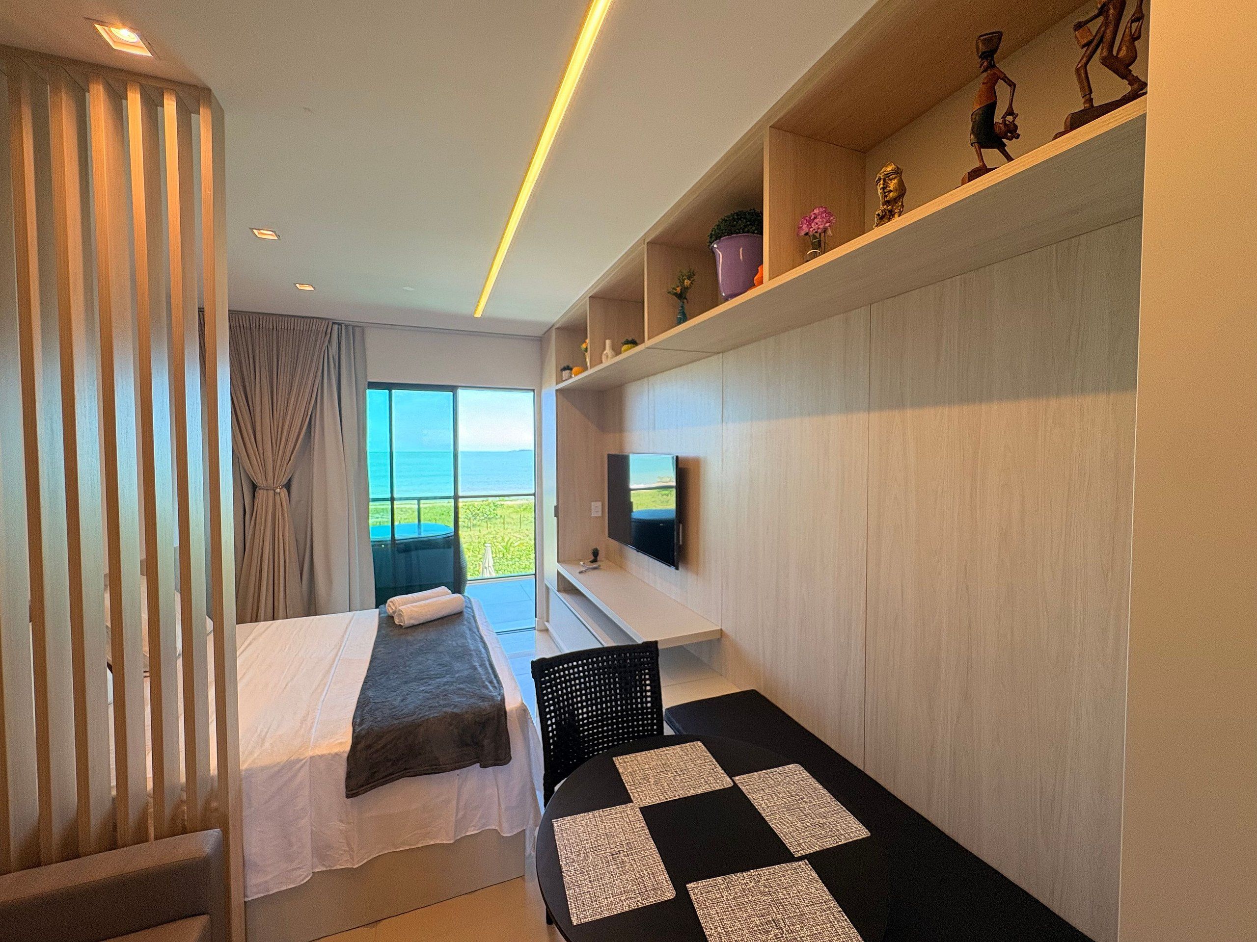 A modern hotel room with a bed, a small seating area, a television, and a view of the sea.