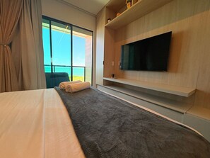Quarto de hotel com cama, televisão de tela plana na parede, uma grande janela com vista e um pequeno móvel com objetos decorativos.