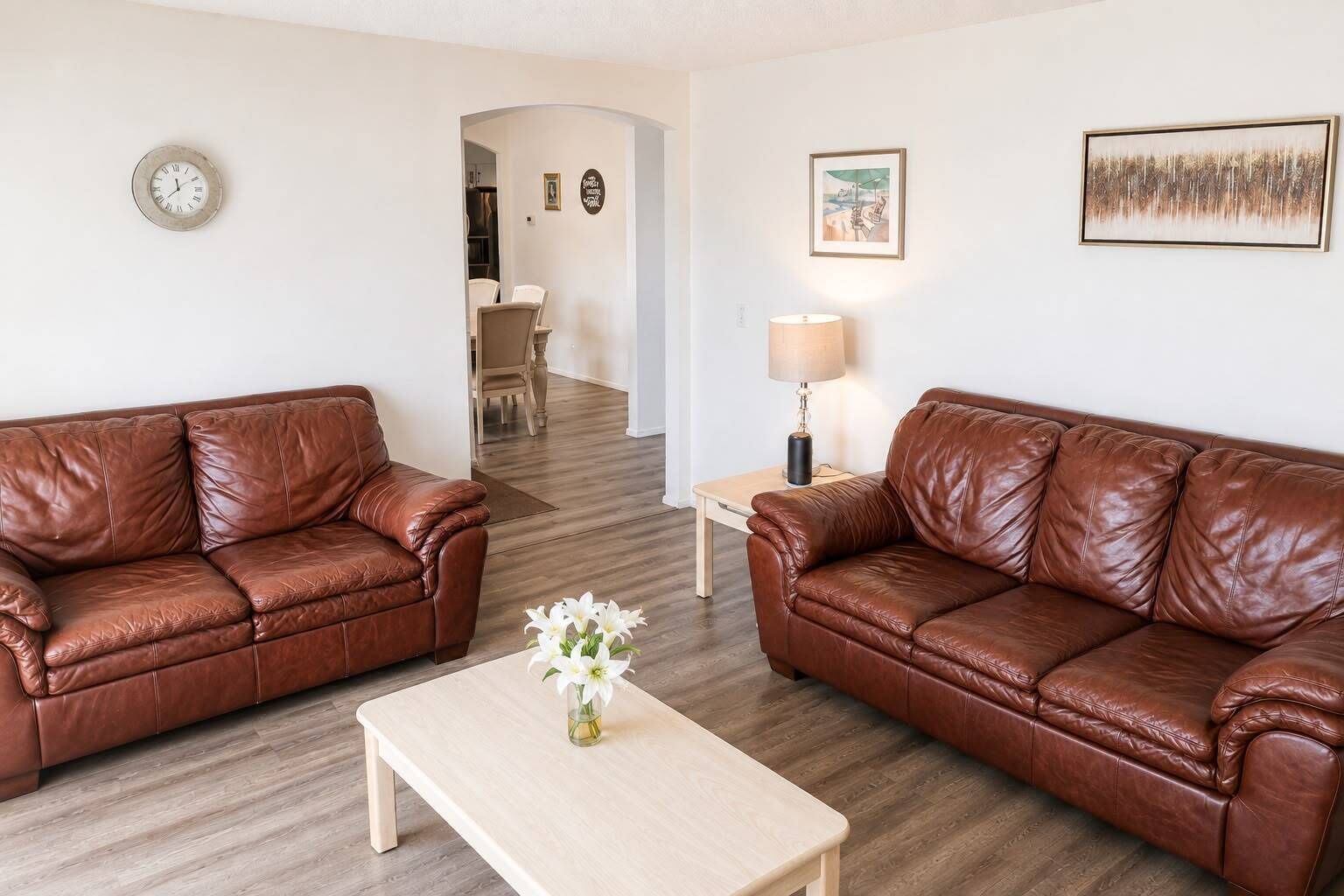 Una sala de estar con dos sofás de cuero marrón, una mesa de centro de madera, una lámpara y un reloj en la pared.