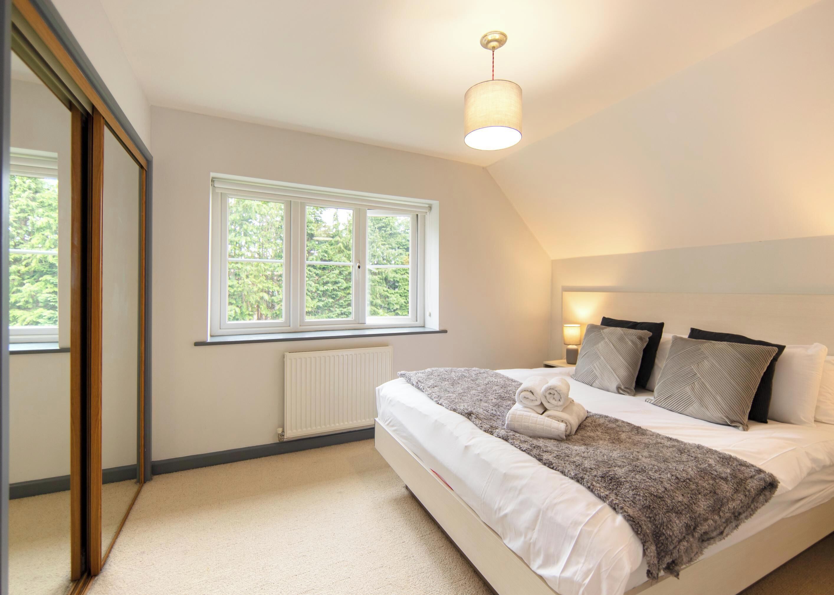 A bedroom with a large window, a bed with pillows and a blanket, a bedside lamp, and a radiator.