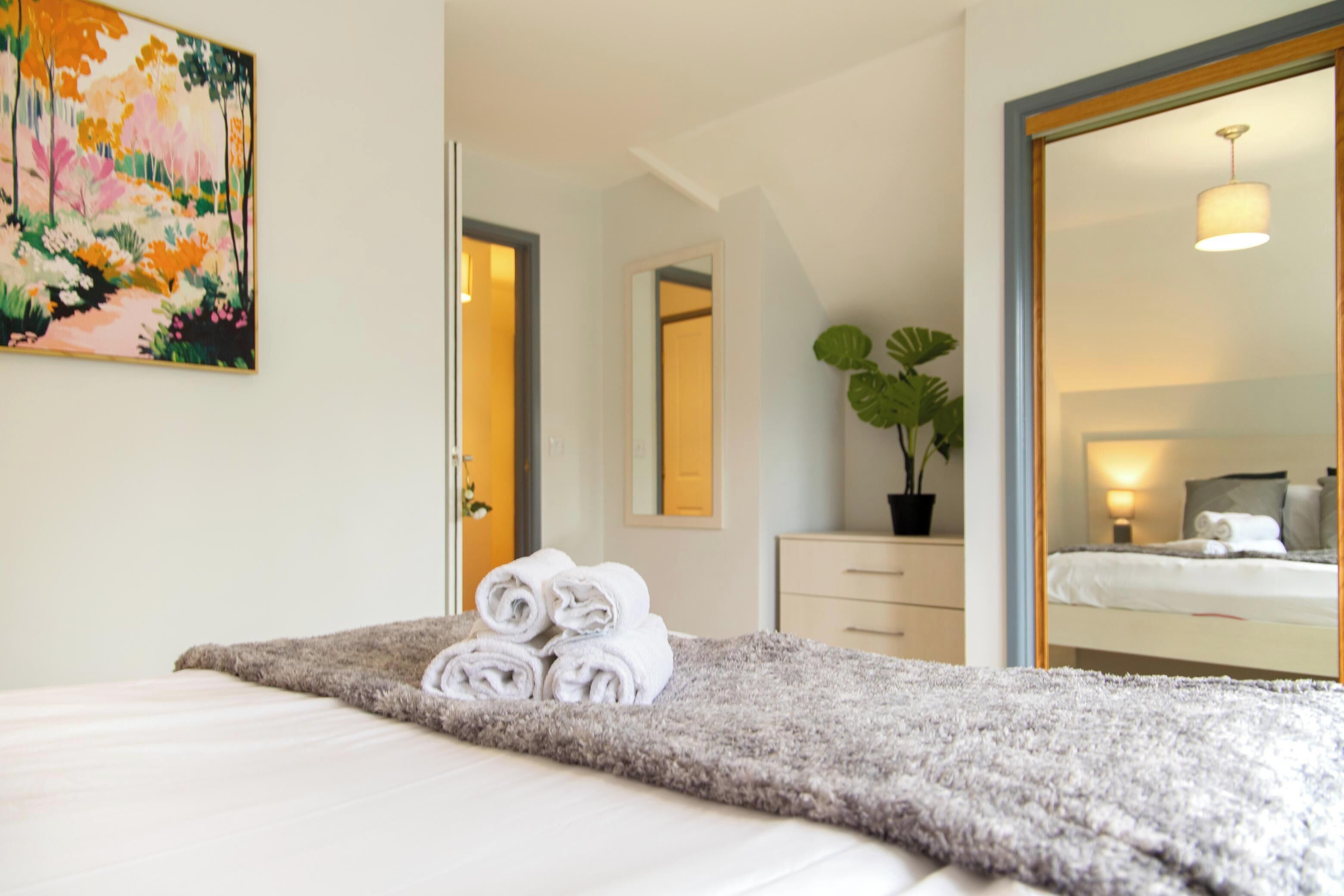 A hotel room with a bed, a mirror, a potted plant, and a painting on the wall.