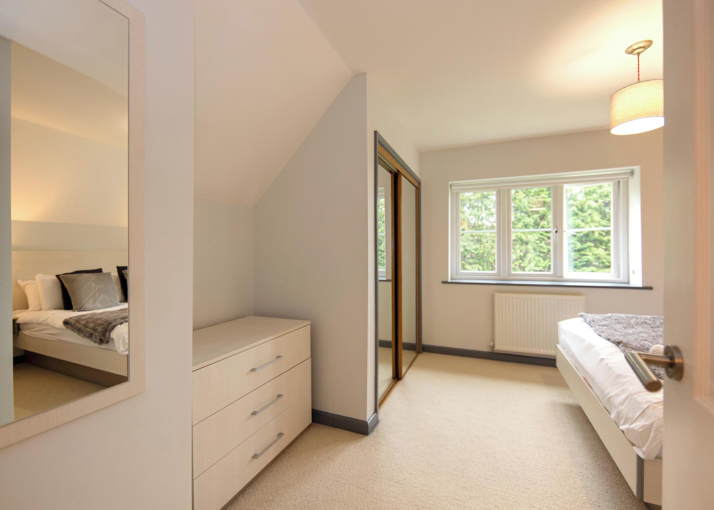 Ein Schlafzimmer mit einem Bett, einer Kommode, einem Spiegel, einem Fenster mit Blick ins Grüne und einer Heizung.