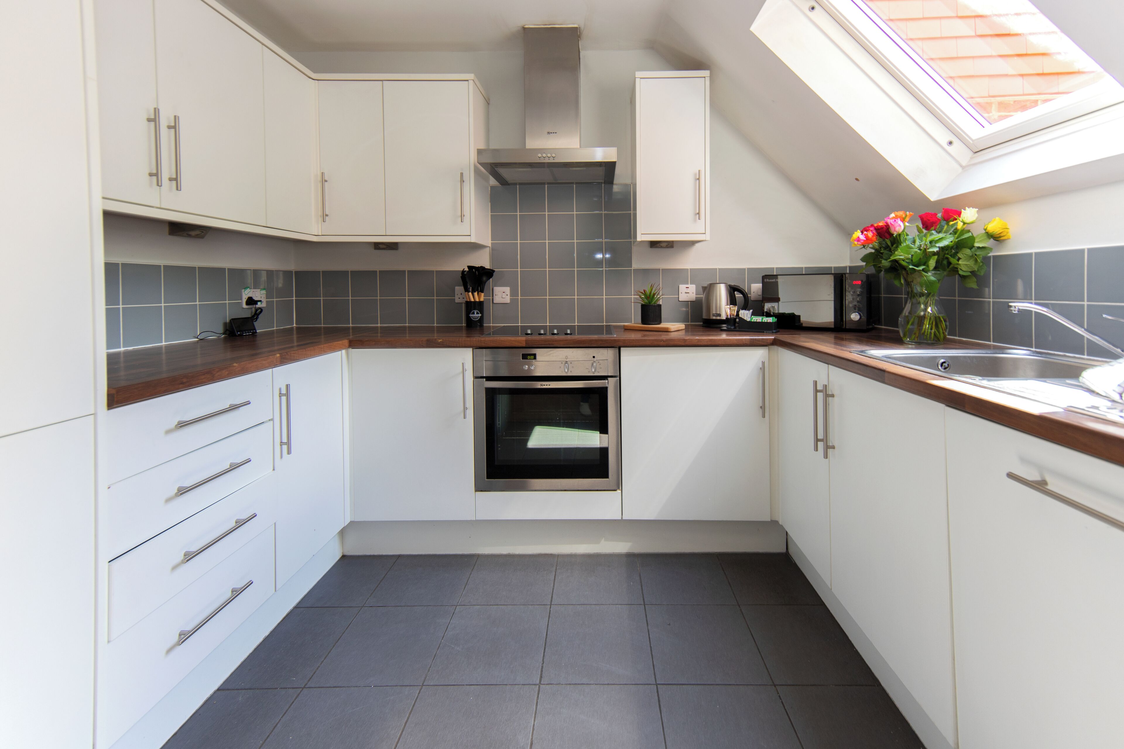 A modern kitchen with white cabinets, a built-in oven, a coffee machine, and a skylight.
