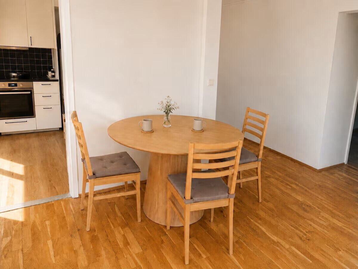A dining area with a round wooden table and four chairs, a vase with flowers, and a kitchen in the background.