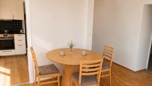 Un comedor con una mesa redonda de madera y cuatro sillas, un jarrón con flores, y una cocina al fondo.
