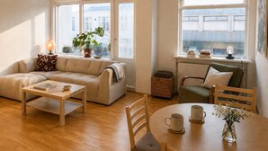 Sala de estar con sofá, mesa de centro y mesa de comedor con sillas. Hay ventanas con cortinas, una lámpara y una planta.