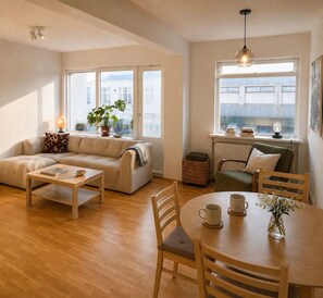 Ein Wohnzimmer mit einer Couch, Couchtisch und Esstisch mit Stühlen. Es gibt Fenster mit Vorhängen, eine Lampe und eine Pflanze.