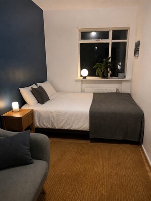 A bedroom with a bed, bedside table, lamp, and a window with a view of the outside. - Hallgrímskirkja View – Central 2-Bedroom (Reykjavíkurborg)