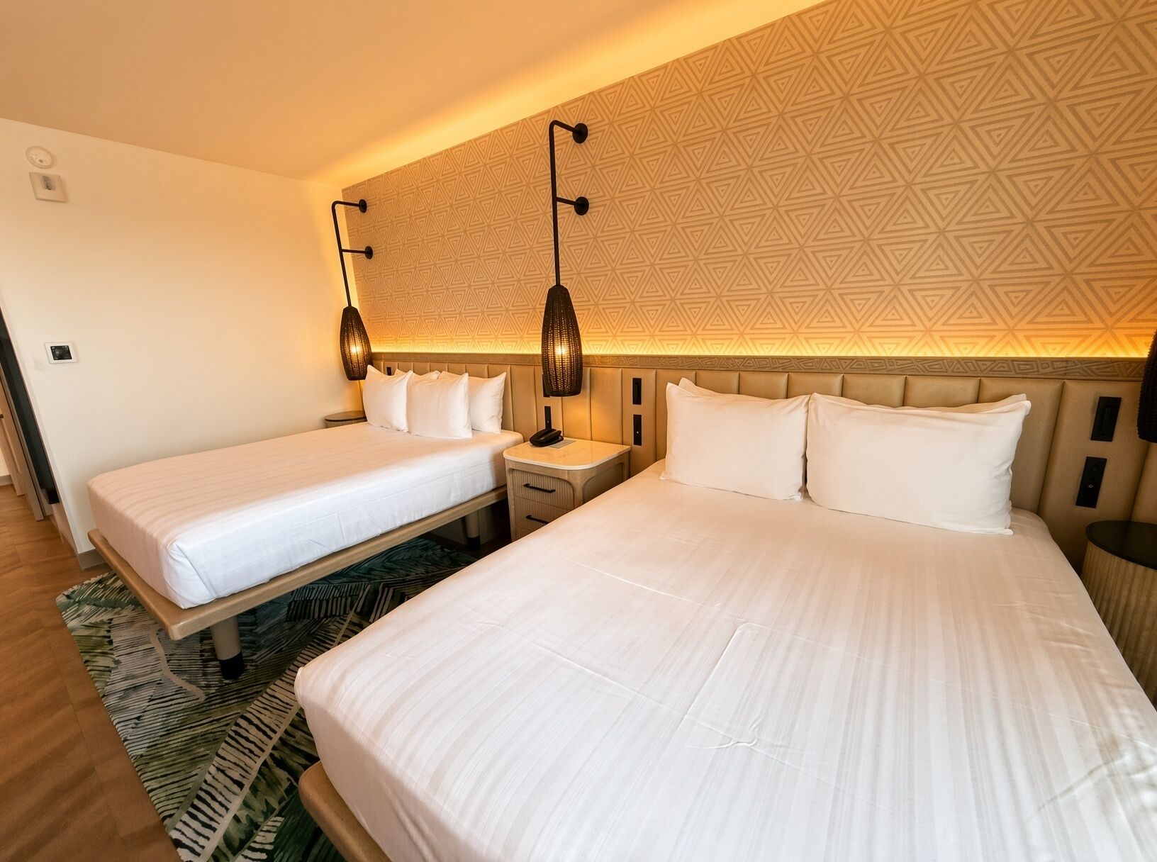 A hotel room with two beds, a patterned wall, and a nightstand with a lamp.