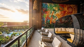Un balcón con vista a una piscina y palmeras, un mural y un sofá de mimbre.