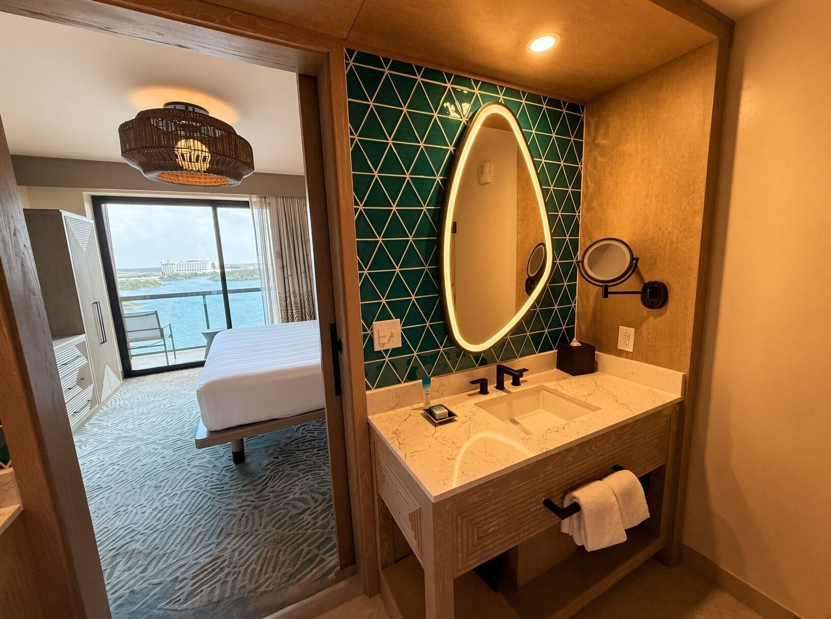 A hotel bathroom with a large oval mirror, a double sink, and a view of a balcony.