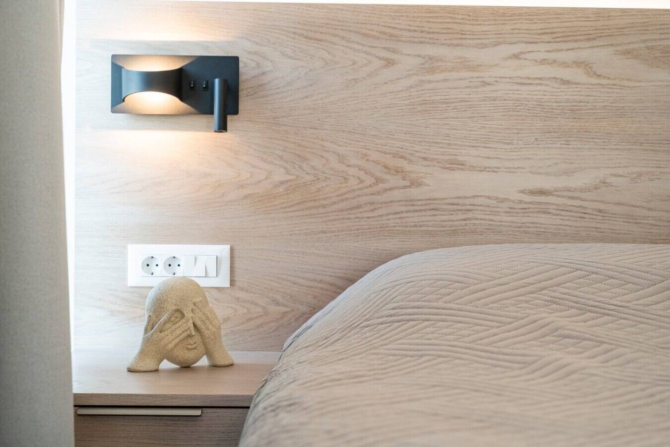 Un letto rifatto con un copriletto a trama decorativa, un lampadario fissato al muro e una presa di corrente su un comodino in legno.