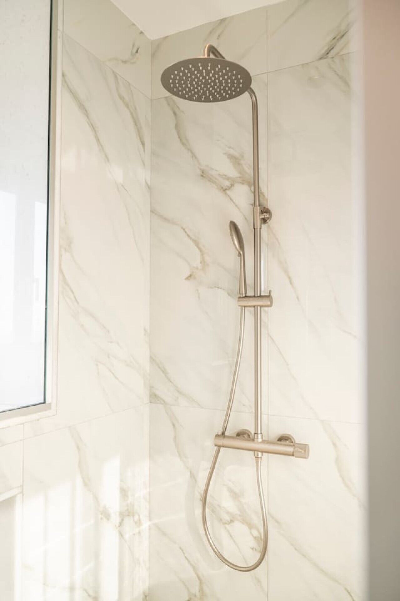 A modern shower with a handheld showerhead and a handheld faucet against a marble wall.