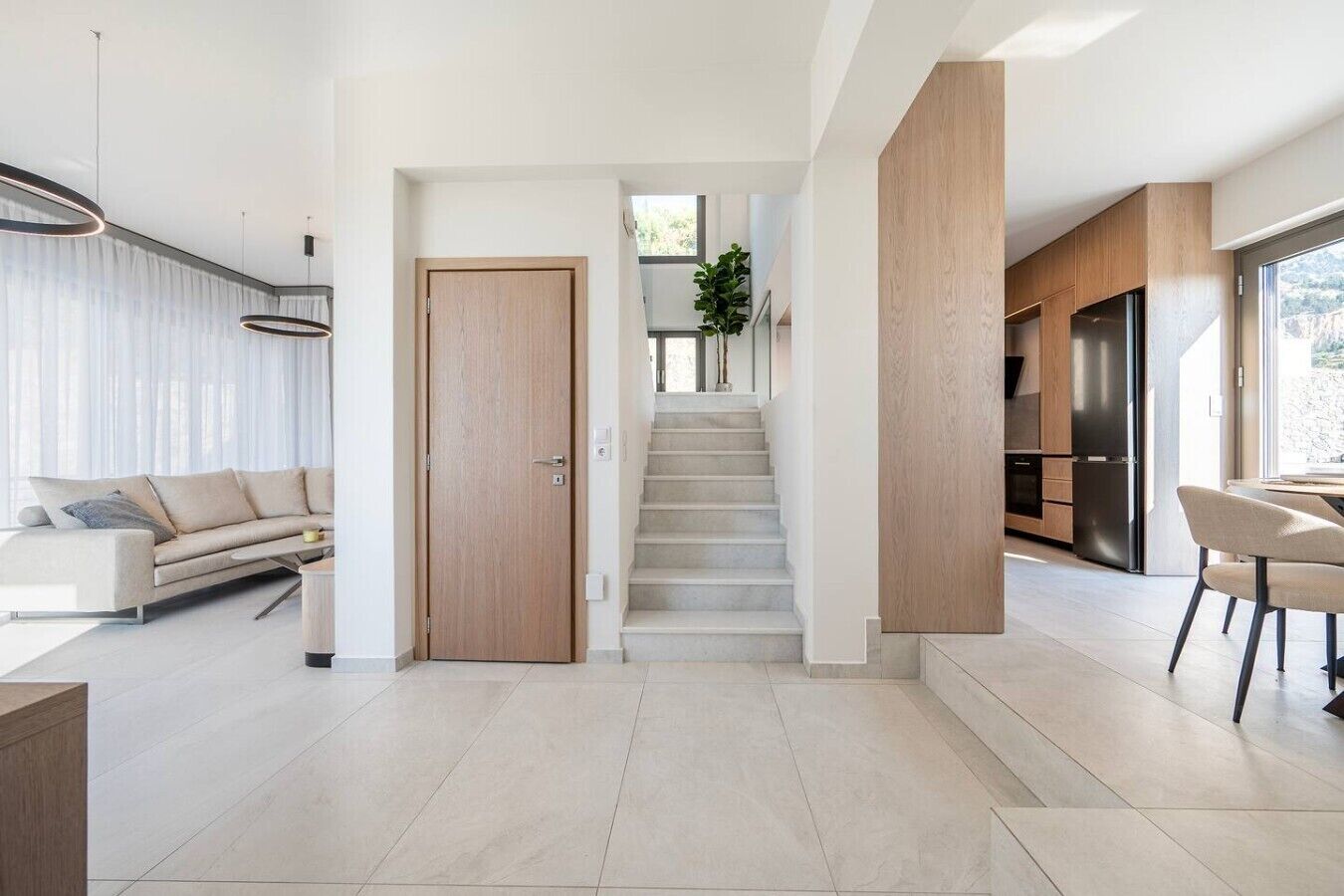 A modern interior with a staircase leading to a second floor, a dining area with chairs, and a kitchen with appliances.