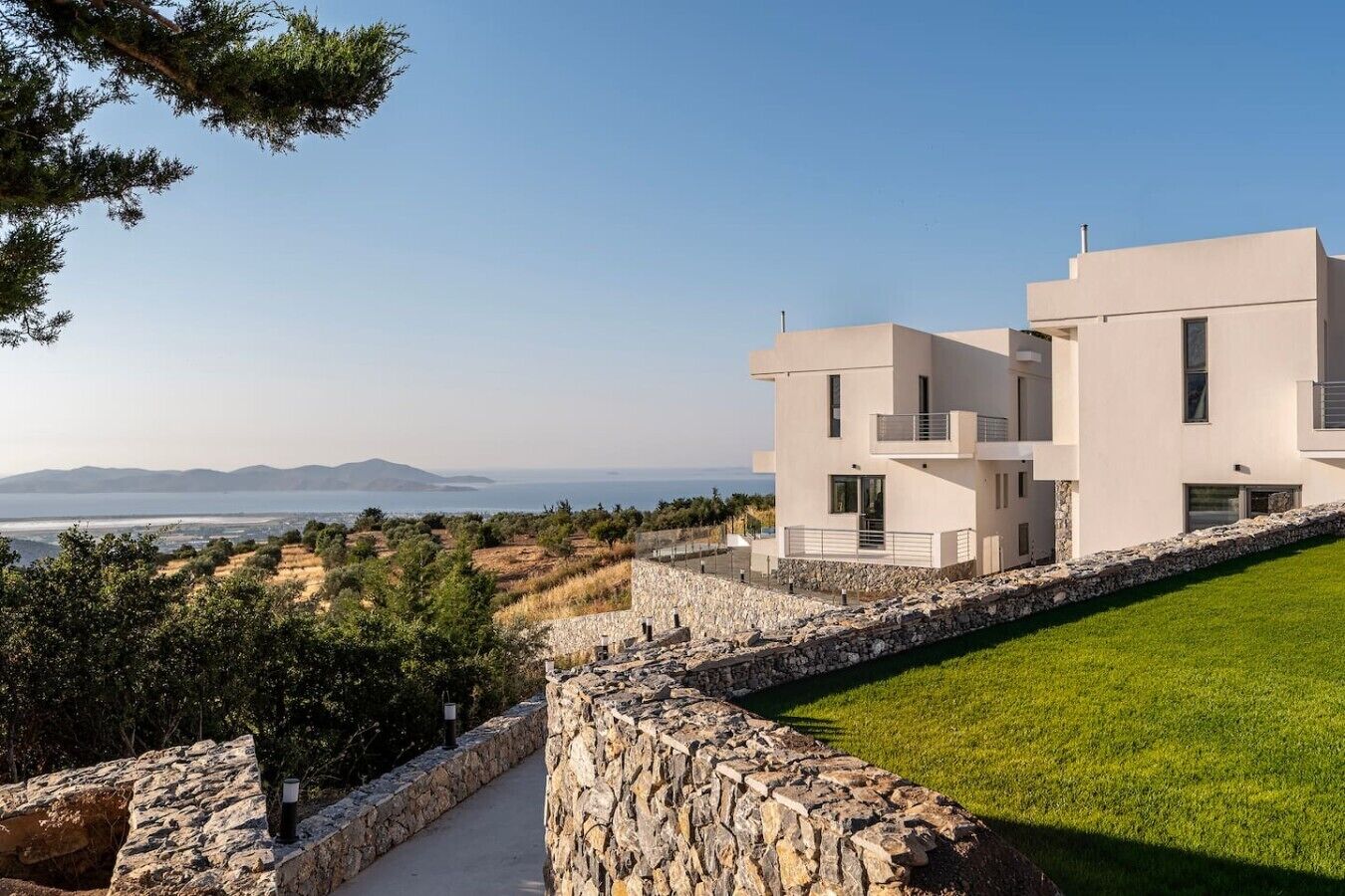 Un bâtiment moderne à deux étages, doté d’un mur de soutènement en pierre, d’une pelouse bien entretenue et offrant une vue sur la mer et les montagnes lointaines.