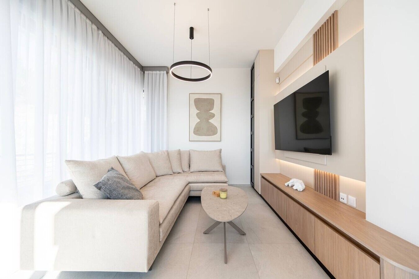 A modern living room with a sectional sofa, a round coffee table, a flat-screen TV mounted on the wall, and a pendant light.