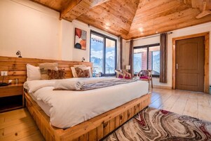 Un dormitorio con estructura de madera, una cama grande, una mesita de noche, una silla y vistas a las montañas a través de las ventanas.