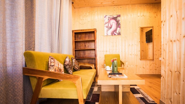 A cozy room with a yellow sofa, a wooden bookshelf, and a table with a vase. - Aaryam Resort Auli (Joshimath)
