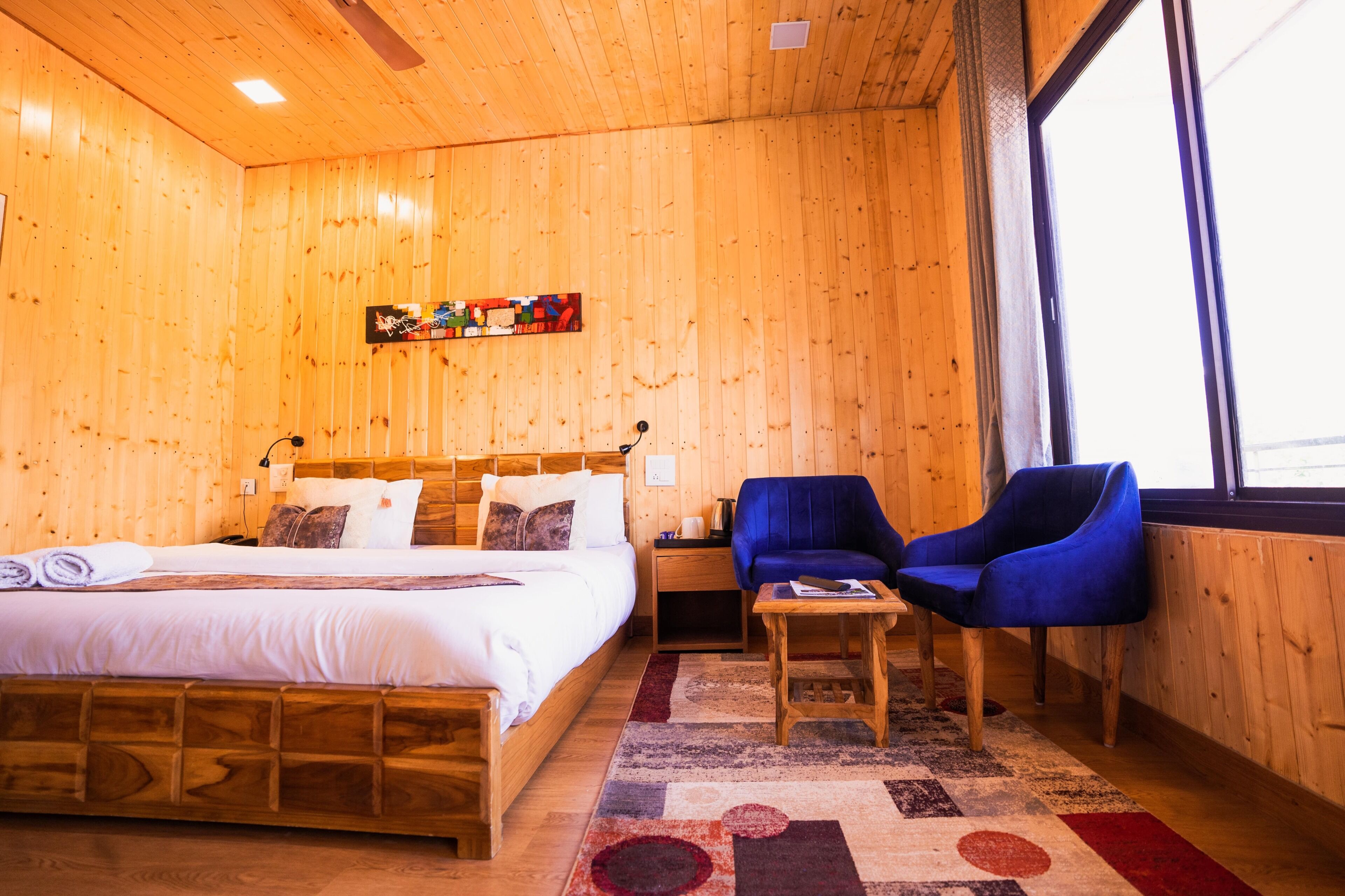 A wooden room with a bed, a blue chair, a small table, and a window.
