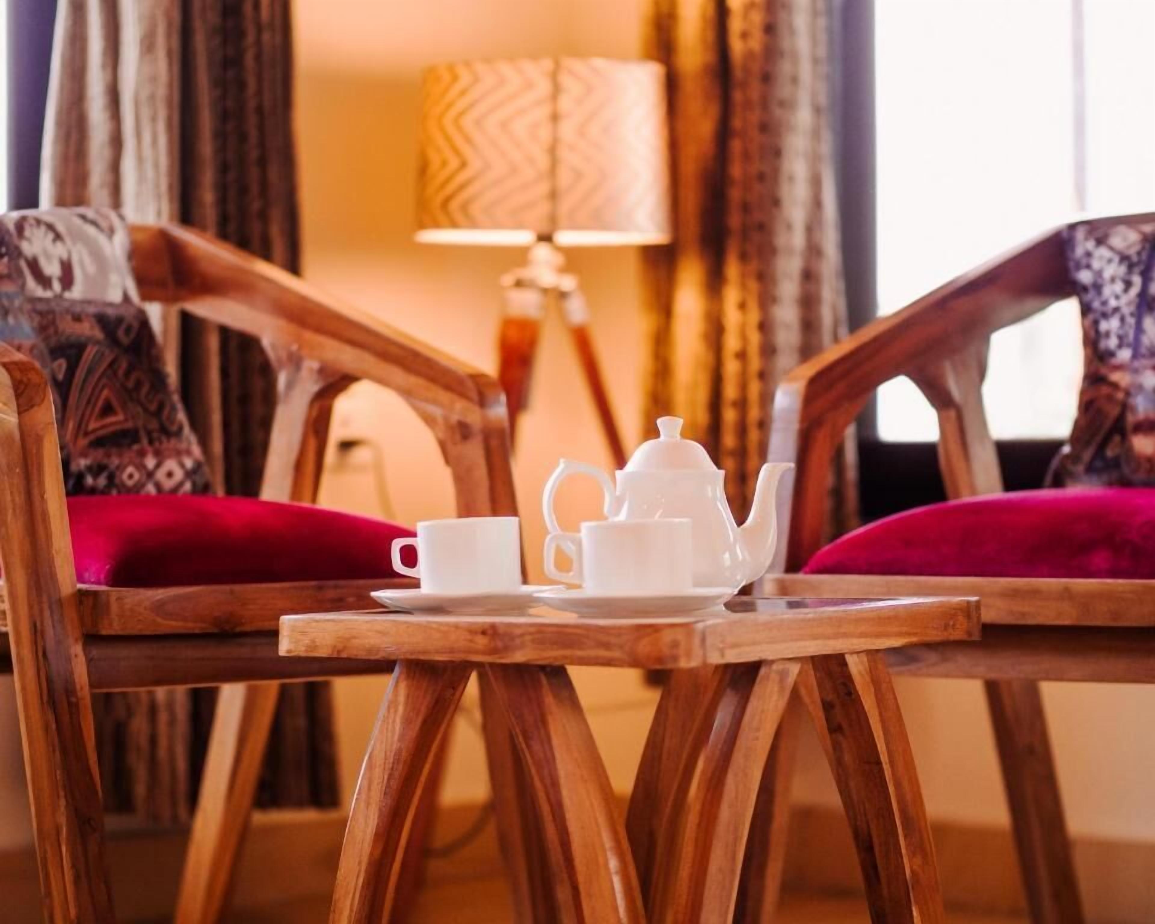 Une table en bois, sur laquelle sont posés une théière et une tasse, est placée entre deux chaises dotées de coussins rouges.