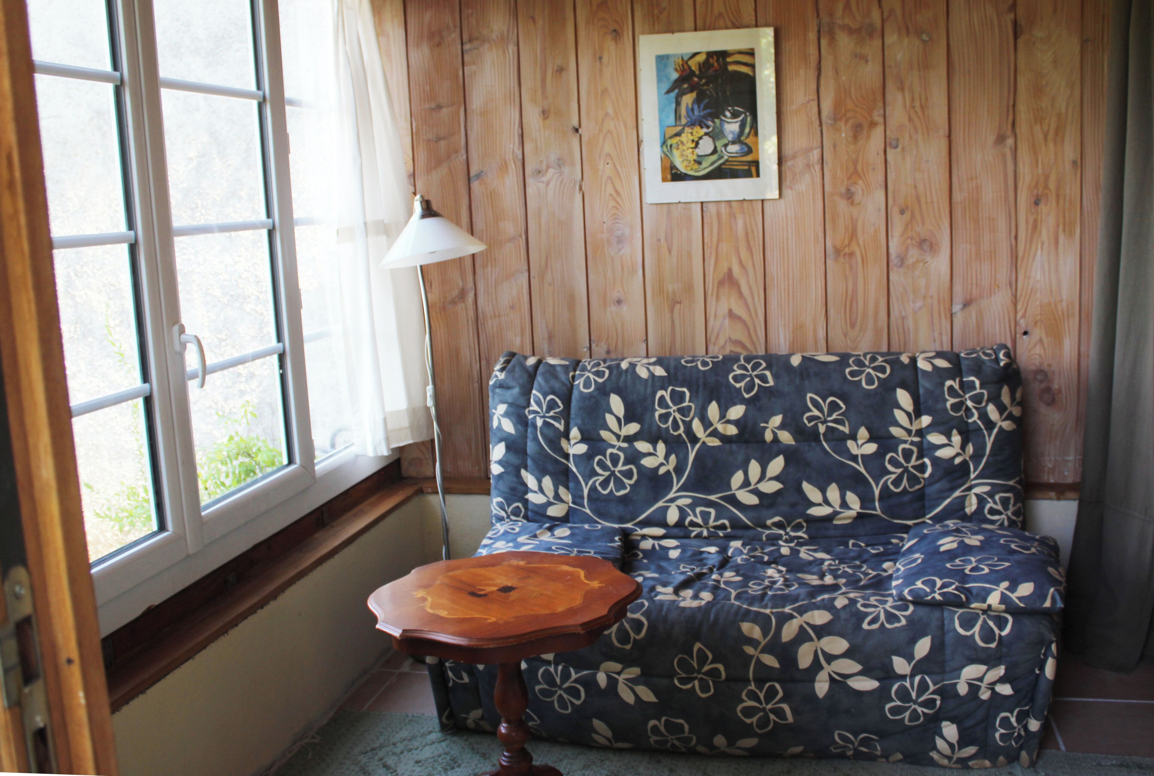 A room with a wooden wall, a window, a sofa with a floral pattern, and a small wooden table.