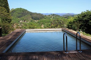 Eine Holzterrasse führt zu einem rechteckigen Pool mit Metallgeländern, umgeben von üppiger Vegetation und einer Bergkulisse.