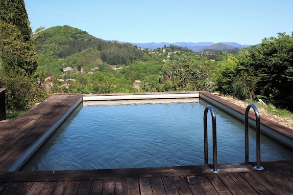 Eine Holzterrasse führt zu einem rechteckigen Pool mit Metallgeländern, umgeben von üppiger Vegetation und einer Bergkulisse.