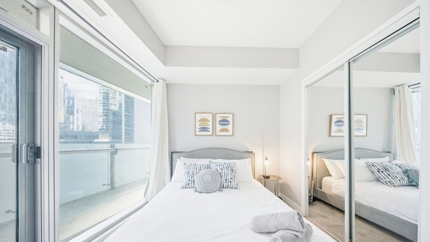 A hotel room with a large bed, a bedside table with a lamp, a window with a view of buildings, and a sliding door. - Founder's Club York - 3 Bedroom Condo  (Toronto)