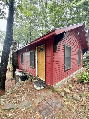 Eine kleine, eingeschossige Holzhütte mit roter Fassade, einer Holztür und einem Steinkamin.