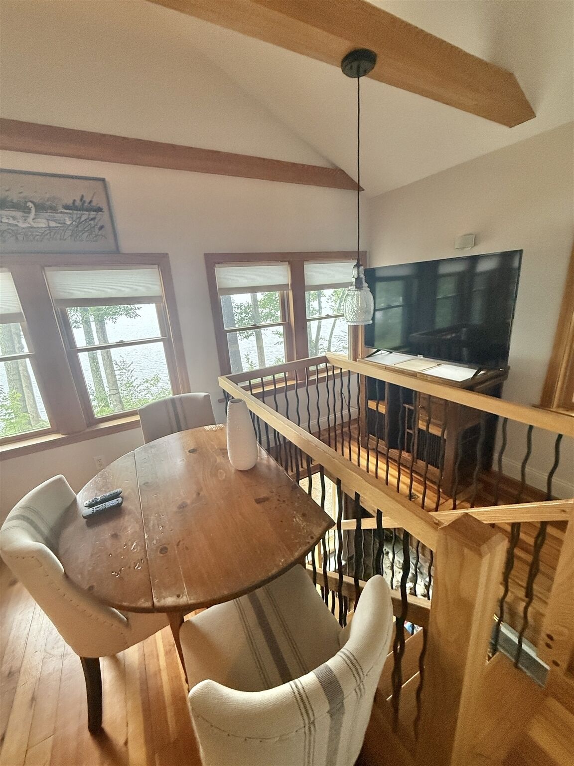 Een eethoek met een houten tafel en stoelen, een televisie op een standaard, en uitzicht op bomen door de ramen.