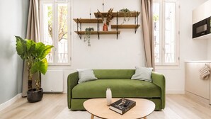 En grøn sofa med to puder, et rundt sofabord med en bog, en potteplante og en vægmonteret reol med pynteting.