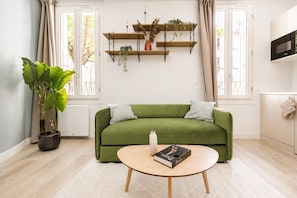 Ein grünes Sofa mit zwei Kissen, ein runder Couchtisch mit einem Buch, eine Topfpflanze und ein an der Wand befestigtes Regal mit Dekoration.