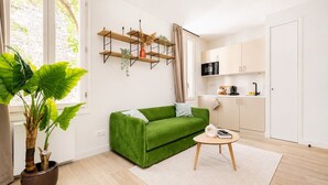 En moderne stue med en grøn sofa, et rundt bord, en potteplante og et tekøkken med skabe og en mikrobølgeovn.