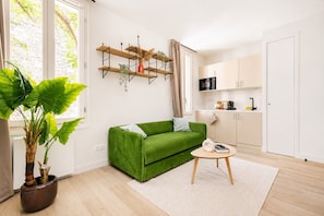 A modern living room with a green sofa, a small round table, a potted plant, and a kitchen area with cabinets and a microwave.