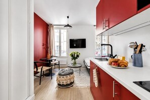 Eine moderne Küche mit roten Schränken, ein Essbereich mit rundem Tisch und Stühlen, ein kleiner Teppich und ein an der Wand befestigter Fernseher.
