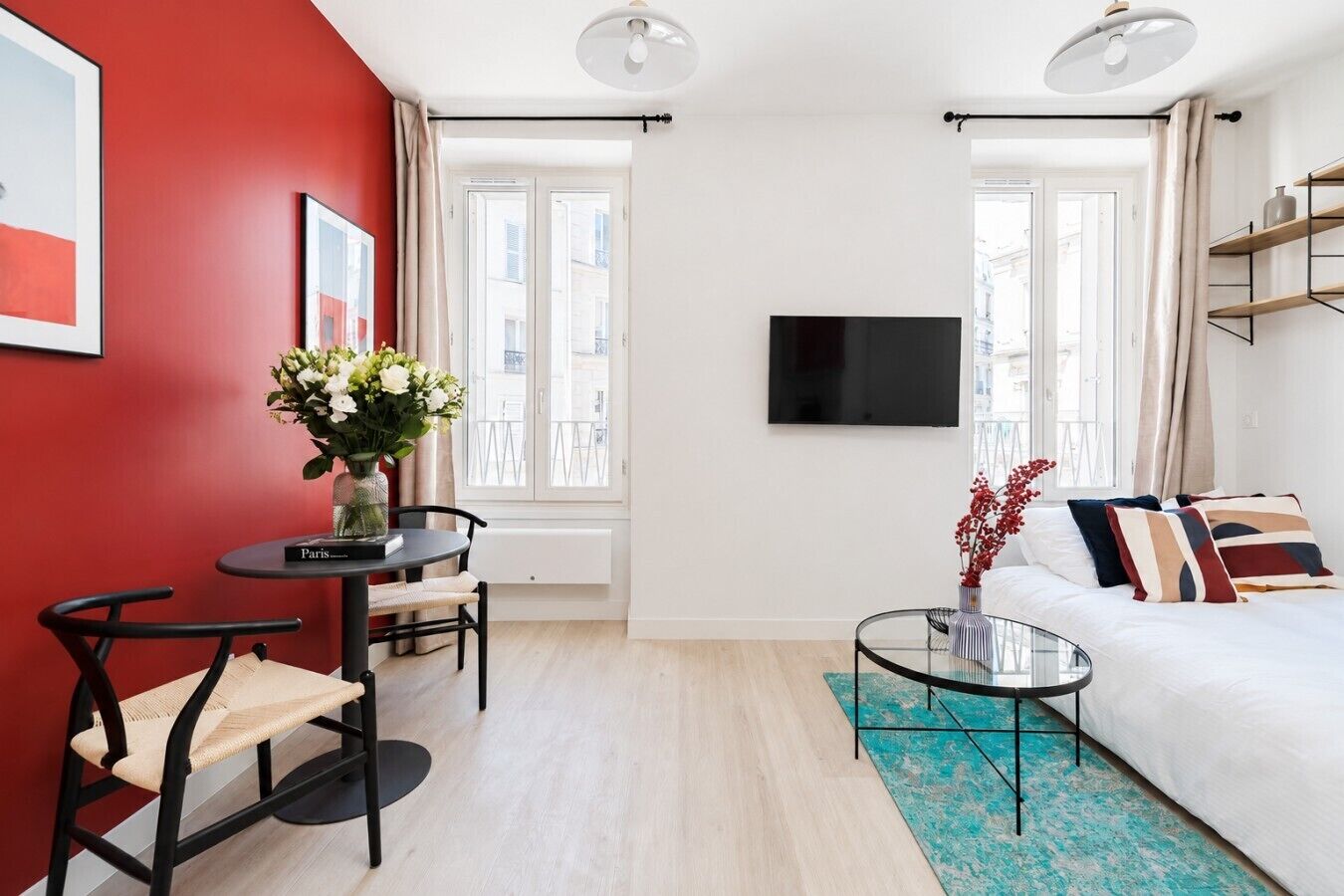 A modern hotel room with a red accent wall, a round dining table with chairs, a sofa, a TV mounted on the wall, and a window with curtains.