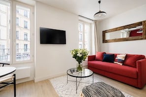 A modern living room with a red sofa, a glass coffee table, and a flat-screen TV mounted on the wall. - Lavie Maison : Charming Studio near to Montmartre (Paris)