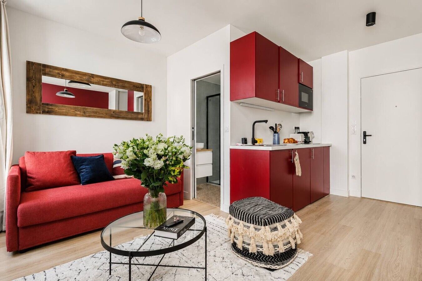 Ein kompakter Wohnraum mit einem roten Sofa, einem runden Couchtisch, einem kleinen Hocker, einer Küchenzeile mit roten Schränken und einer weißen Tür.