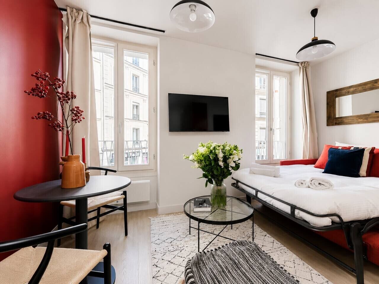 Una habitación de hotel moderna con una cama, un televisor, una mesita con un florero y un ventanal con cortinas.