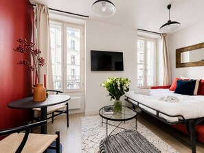 A modern hotel room with a bed, a TV, a small table with a vase, and a window with curtains. - Lavie Maison : Charming Studio near to Montmartre (Paris)