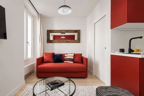 A modern living room with a red sofa, a glass coffee table, and a kitchen area with a red cabinet and a black faucet. - Lavie Maison : Charming Studio near to Montmartre (Paris)