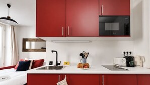 A compact kitchen with red cabinets, a black microwave, and a small countertop with a toaster and coffee maker. - Lavie Maison : Charming Studio near to Montmartre (Paris)