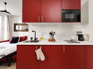 A compact kitchen with red cabinets, a black microwave, and a small countertop with a toaster and coffee maker. - Lavie Maison : Charming Studio near to Montmartre (Paris)