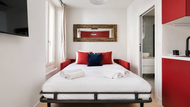 A modern hotel room with a bed, a flat-screen TV mounted on the wall, a wooden-framed mirror, and a red accent wall. - Lavie Maison : Charming Studio near to Montmartre (Paris)
